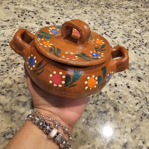 May include: A small, brown clay pot with a lid. The pot is decorated with green leaves, orange and red flowers, and white dots. The lid has a handle and is also decorated with green leaves and flowers.