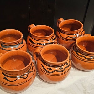May include: Six terracotta mugs with handles, each adorned with a black painted design. The mugs have a rustic, handmade appearance and are arranged on a white surface. The mugs are approximately 4 inches in diameter.