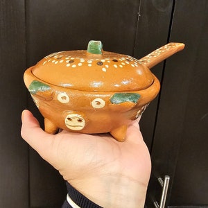 May include: Handmade terracotta pottery bowl with lid. The bowl is a warm orange color with white polka dots and green accents. It has a handle and three small feet. The lid has a green knob.