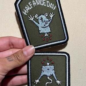 Puede incluir: Dos parches bordados verdes con bordes blancos. El parche superior presenta un monstruo de dibujos animados con el texto "Half a Nice Day". El parche inferior presenta un monstruo de dibujos animados con el texto "It Could Be Worse".