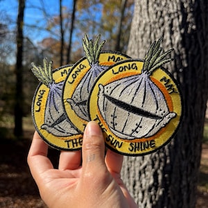 以下が含まれることがあります： 黄色い背景に「Make Long The Sun Shine」という文字が書かれた、漫画の玉ねぎが描かれた刺繍パッチが3つあります