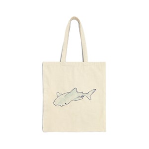 Pode incluir: Uma bolsa tote de lona natural com uma ilustração azul e verde de um tubarão na frente.