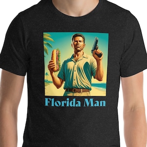 Puede incluir: Una camiseta negra con un gráfico de un hombre sosteniendo un sándwich y una pistola. El gráfico está situado en una escena de playa con palmeras. El texto "Florida Man" está impreso debajo del gráfico.