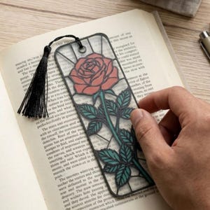 May include: A rectangular bookmark featuring a stained glass style design of a pink rose with green leaves and a stem. The bookmark has a black outline, a metal eyelet at the top, and a black tassel attached with a thin black cord.