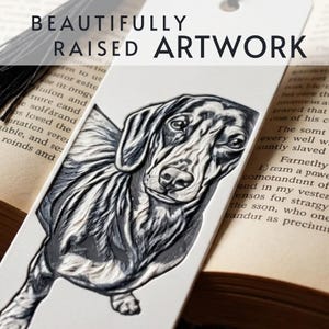 May include: A white bookmark featuring a black and white raised illustration of a dachshund dog. The bookmark includes a black tassel at the top and the text BEAUTIFULLY RAISED ARTWORK printed in black across the upper portion of the bookmark.
