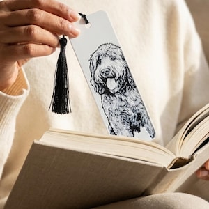 May include: A white rectangular bookmark featuring a detailed black and white ink illustration of a curly-haired dog with its tongue out. The bookmark has a hole at the top with a black thread tassel attached, resting inside an open book.