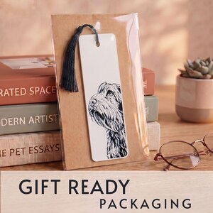 May include: A rectangular white bookmark featuring a black and white ink illustration of a dog's head and neck. The bookmark has a hole at the top with a black tassel attached and is enclosed in a clear plastic sleeve against a brown card backing.