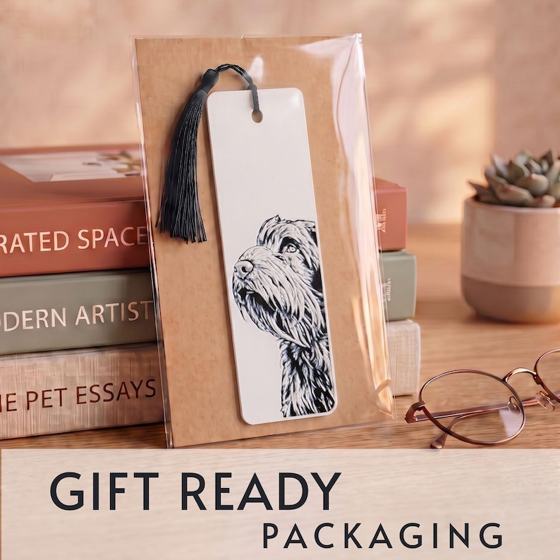 May include: A rectangular white bookmark featuring a black and white ink illustration of a dog's head and neck. The bookmark has a hole at the top with a black tassel attached and is enclosed in a clear plastic sleeve against a brown card backing.