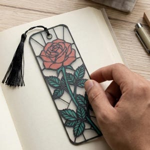 May include: A rectangular bookmark featuring a black frame with a stained glass style design of a pink rose with green leaves and a stem. The bookmark includes a black tassel attached to the top through a small metal eyelet, resting on an open book.
