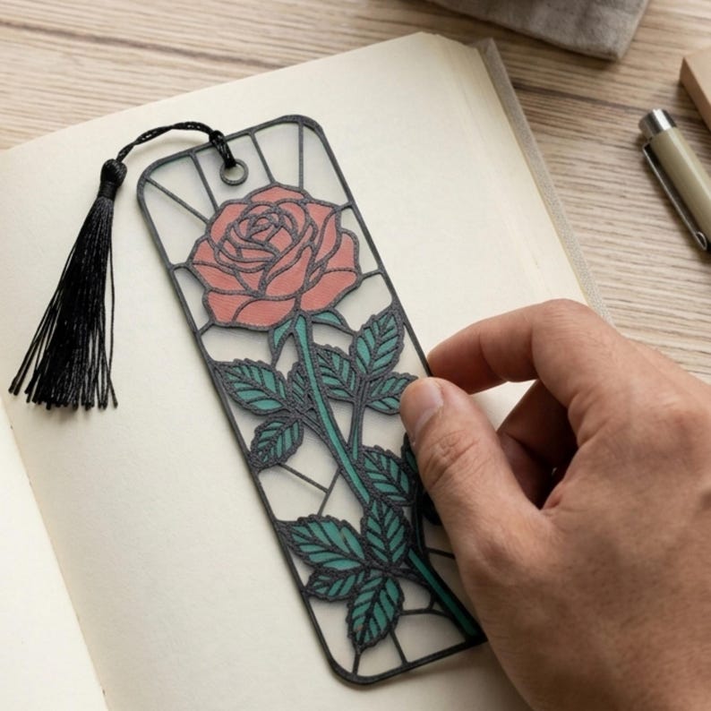 May include: A rectangular bookmark featuring a black frame with a stained glass style design of a pink rose with green leaves and a stem. The bookmark includes a black tassel attached to the top through a small metal eyelet, resting on an open book.