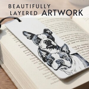 May include: A white rectangular bookmark featuring a black and white illustration of two Boston Terrier dogs stacked vertically. The bookmark has a black cord attached at the top and is resting on an open book with printed text visible underneath.
