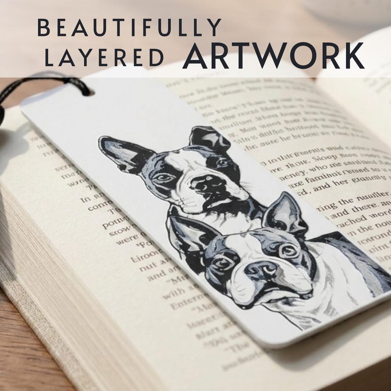 May include: A white rectangular bookmark featuring a black and white illustration of two Boston Terrier dogs stacked vertically. The bookmark has a black cord attached at the top and is resting on an open book with printed text visible underneath.