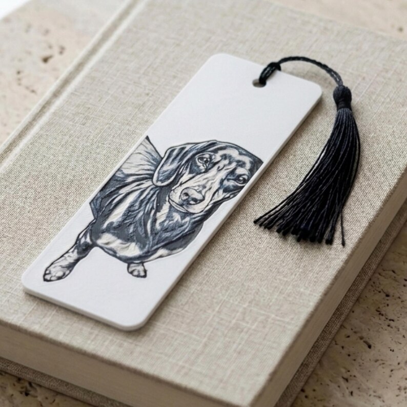 May include: A rectangular white bookmark featuring a detailed black and white ink illustration of a dachshund dog. The bookmark has a hole at the top with a black thread tassel attached, providing a decorative accent to the printed canine portrait.