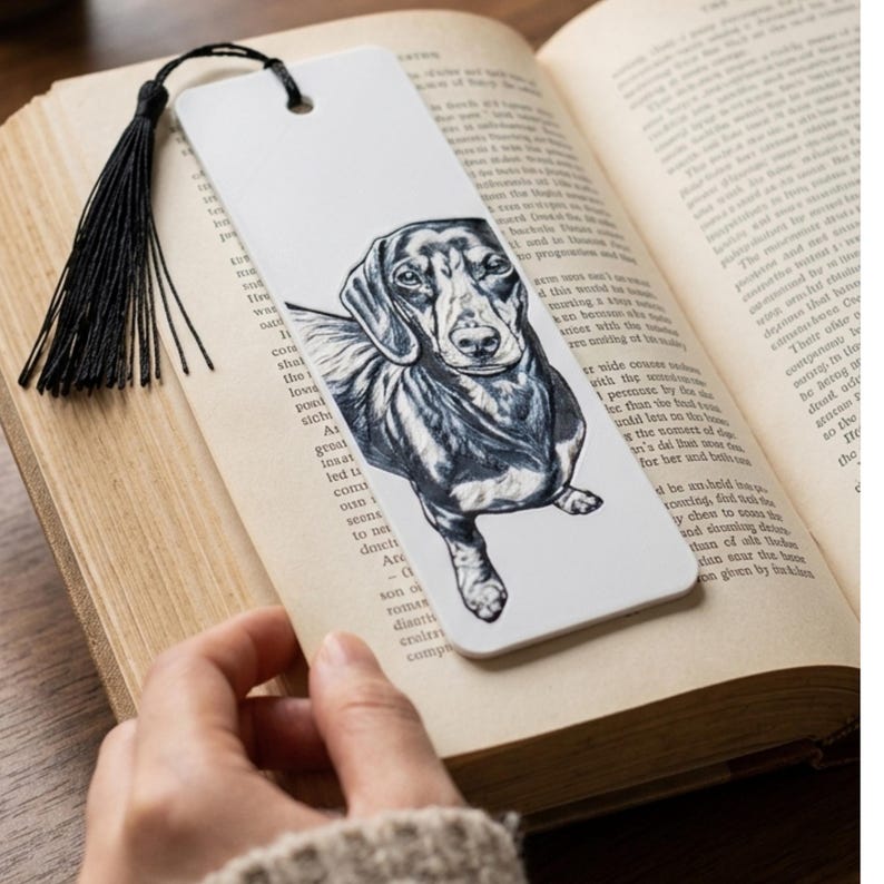 May include: A white rectangular bookmark featuring a detailed black and white ink illustration of a dachshund dog. The bookmark has a hole at the top with a black thread tassel attached, resting on the open pages of a printed book.