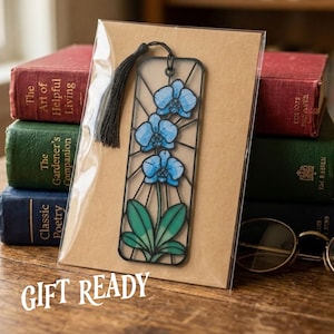 May include: A rectangular bookmark featuring a stained-glass style illustration of a blue orchid with green leaves, enclosed in a black frame with a black tassel at the top, packaged in a clear plastic sleeve on a brown card backing.