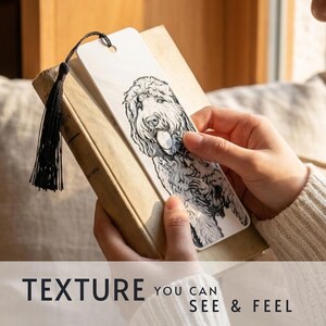 May include: A rectangular white bookmark featuring a detailed black and white ink-style illustration of a shaggy dog with its tongue out. The bookmark has a hole at the top with a black tassel attached and is placed inside a closed hardcover book.