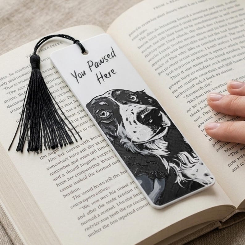 May include: A white rectangular bookmark featuring a black and white illustration of a dog's face and the text "You Paused Here" in black script. The bookmark includes a black string tassel attached through a hole at the top edge.