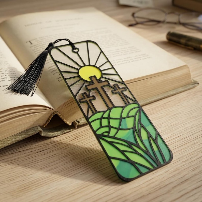 May include: A rectangular bookmark featuring a black frame with a stained glass style design of three brown crosses on a green hill under a yellow sun. The bookmark includes a black tassel attached to the top and has a semi-transparent appearance.