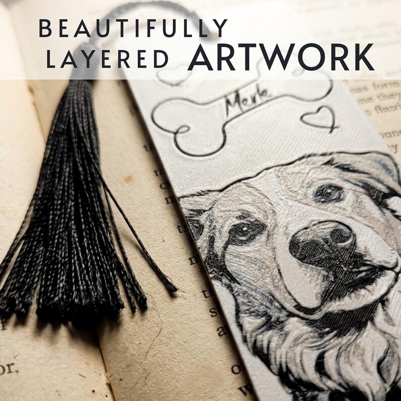 May include: A rectangular bookmark featuring a detailed engraved portrait of a dog, a bone shape with the name Merle written inside, and small heart outlines. The bookmark is topped with a black thread tassel and has a textured white surface.