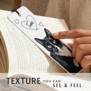 May include: A white rectangular bookmark featuring a black and white illustration of a cat with a heart outline containing the text Tuna and two smaller hearts. The bookmark has a black string loop at the top and is placed inside an open book.