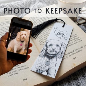 May include: A rectangular white paper bookmark featuring a black ink illustration of a curly-haired dog with a collar and tag. The name Daisy is written inside a heart at the top, and a black tassel is attached to the top hole of the bookmark.