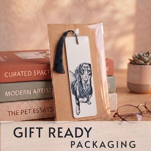 May include: A rectangular white bookmark featuring a black and white ink illustration of a dachshund dog. The bookmark has a black tassel attached to the top and is enclosed in a clear plastic sleeve mounted on a brown cardstock backing.