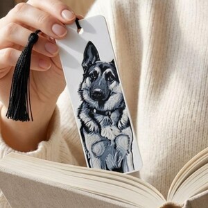 May include: A rectangular white bookmark featuring a detailed black, gray, and white illustration of a German Shepherd dog. The bookmark has a hole at the top with a black thread tassel attached, showcasing a portrait of the canine facing forward.