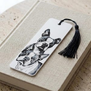 May include: A rectangular white bookmark featuring a black and white ink illustration of two Boston Terrier dogs stacked vertically. The bookmark has a hole at the top with a black thread tassel attached, creating a decorative accent for books.