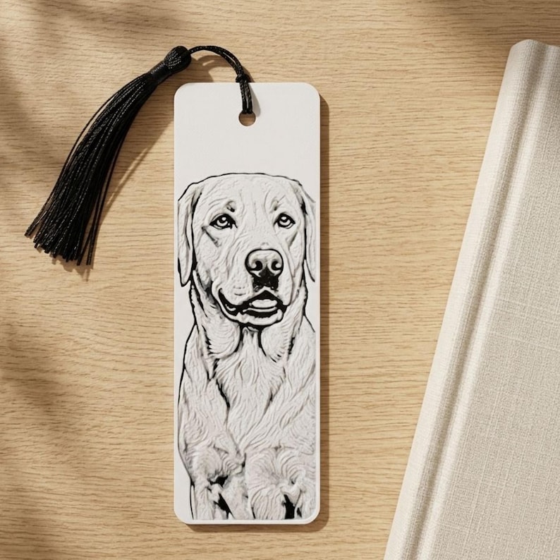 May include: A rectangular white bookmark featuring a detailed black and white line drawing of a Labrador retriever dog. The bookmark has a hole at the top with a black thread tassel attached, providing a decorative accent to the paper accessory.