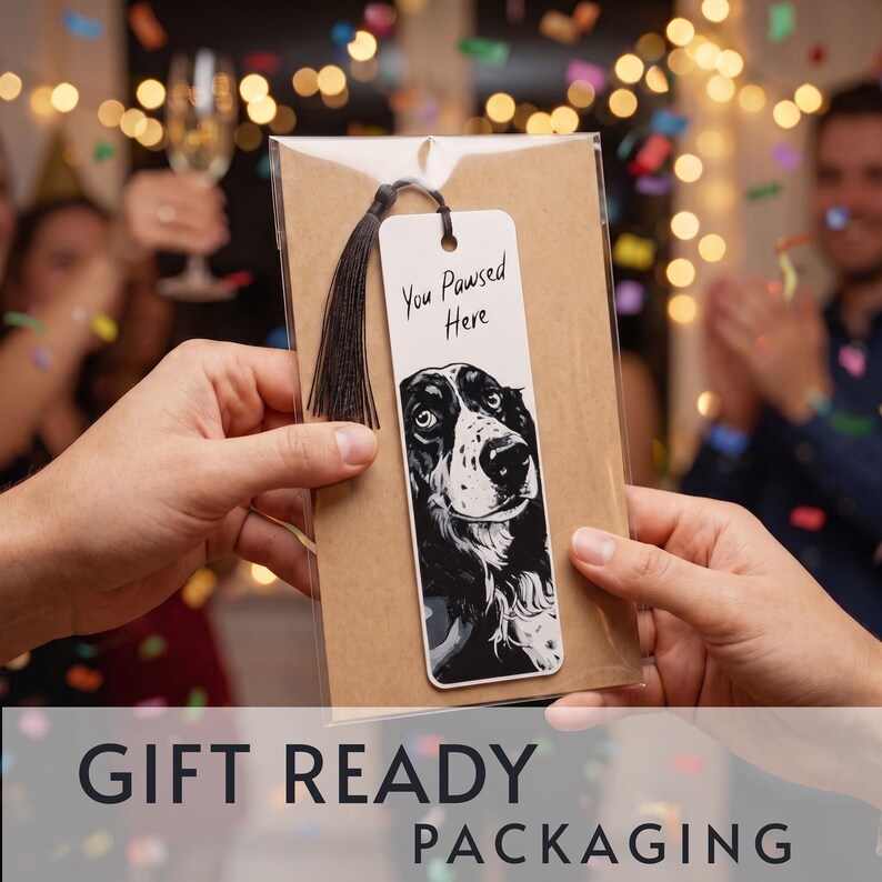May include: A white rectangular bookmark featuring a black and white illustration of a dog's face and the text "You Pawsed Here" in black script. The bookmark has a black tassel attached to the top and is enclosed in clear plastic packaging.