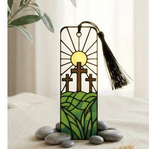 May include: A rectangular metal bookmark featuring a stained-glass style design of three brown crosses on a green hill under a yellow sun with radiating lines. The bookmark has a black cord tassel attached to the top and a circular hole.