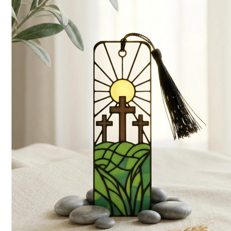May include: A rectangular metal bookmark featuring a stained-glass style design of three brown crosses on a green hill under a yellow sun with radiating lines. The bookmark has a black cord tassel attached to the top and a circular hole.