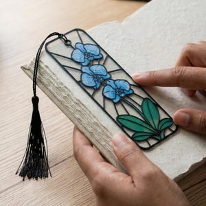 May include: A rectangular bookmark featuring a black frame with a stained glass effect. The design displays three blue orchid flowers with green leaves, connected by thin black lines. A black cord with a matching black tassel is attached to the top.