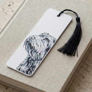 May include: A rectangular white bookmark featuring a detailed black and white ink illustration of a shaggy-haired dog's head. The bookmark has a hole at the top with a black thread tassel attached, providing a contrast to the white surface.