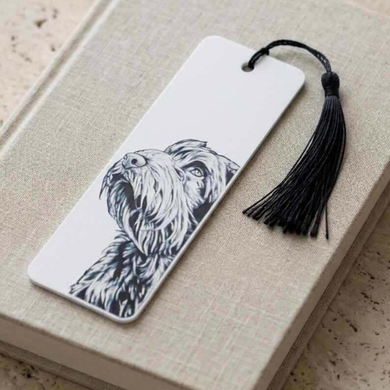 May include: A rectangular white bookmark featuring a detailed black and white ink illustration of a shaggy-haired dog's head. The bookmark has a hole at the top with a black thread tassel attached, providing a contrast to the white surface.