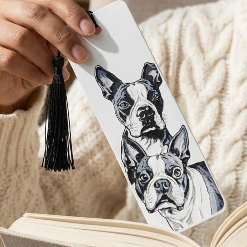 May include: A rectangular white bookmark featuring a detailed black and white illustration of two Boston Terrier dogs stacked vertically. The bookmark is finished with a thin black tassel attached to the top through a small circular hole.