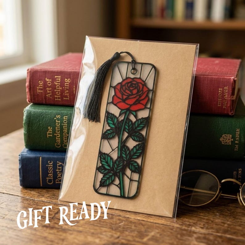 May include: A rectangular bookmark featuring a stained-glass style illustration of a red rose with green leaves and a black outline. The bookmark is encased in a clear plastic sleeve on a tan card and includes a black tassel attached at the top.