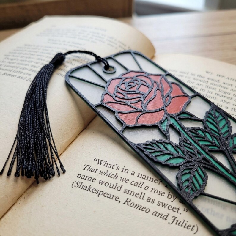 May include: A rectangular bookmark featuring a black frame with a stained-glass style rose design in red and green. The bookmark has a black tassel attached to the top and rests on an open book page with the text What's in a name? That which we call a rose by any other name would smell as sweet.