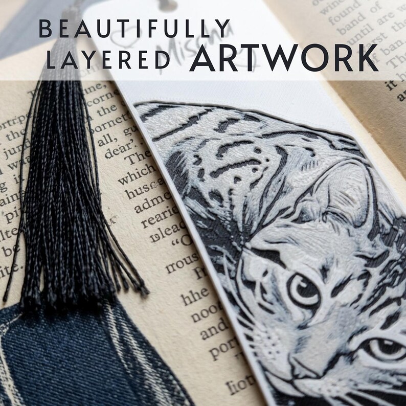 May include: A white bookmark featuring a detailed black and white engraved illustration of a cat's face. The bookmark includes a black tassel attached to the top and displays the text BEAUTIFULLY LAYERED ARTWORK across the upper portion.