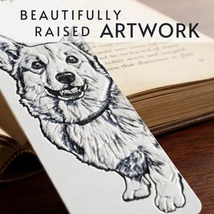 May include: A white rectangular bookmark featuring a raised, black and white illustration of a Corgi dog. The text "BEAUTIFULLY RAISED ARTWORK" is printed in black at the top. The bookmark has a smooth, textured surface with a hole at the top.