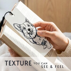 May include: A white rectangular bookmark featuring a detailed black and white ink-style illustration of a Corgi dog. The bookmark has a hole at the top with a black tassel attached and is placed inside an open book with white pages.