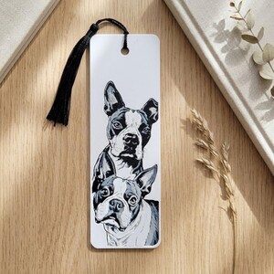 May include: A rectangular white bookmark featuring a black and white illustration of two Boston Terrier dogs stacked vertically. The bookmark has a hole at the top with a black silk tassel attached, providing a decorative accent to the paper design.