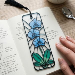 May include: A rectangular bookmark featuring a black frame with a stained glass effect. The design displays three light blue orchid flowers at the top and green leaves at the bottom. A black tassel is attached to the top of the bookmark.