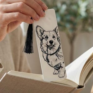 May include: A rectangular white bookmark featuring a detailed black ink illustration of a Corgi dog. The bookmark is made of a smooth material and includes a thin black thread tassel attached to the top through a small circular hole punch.