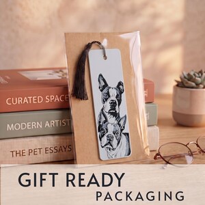 May include: A rectangular bookmark featuring a black and white illustration of two Boston Terrier dogs, one positioned above the other. The bookmark is made of white material, includes a dark brown tassel at the top, and is encased in clear plastic.