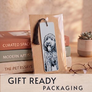 May include: A rectangular bookmark featuring a black and white illustration of a curly-haired dog with its tongue out. The bookmark is made of a light-colored material, includes a dark brown tassel at the top, and is enclosed in clear packaging.