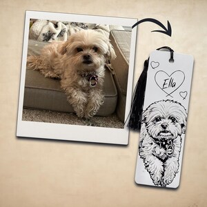 May include: A rectangular white bookmark featuring a black and white line art illustration of a fluffy dog with a collar. The name Ella is written inside a heart shape above the dog, and a black tassel is attached to the top of the bookmark.