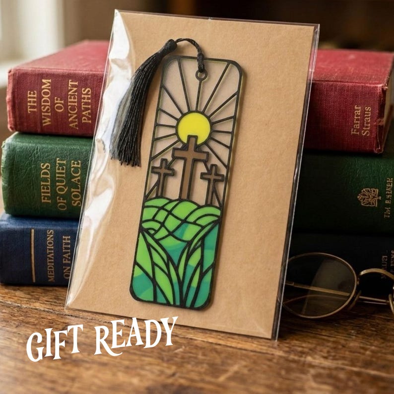 May include: A rectangular bookmark featuring a black frame with a stained glass design of three brown crosses on a green hill under a yellow sun. A black tassel is attached to the top. The bookmark is inside a clear sleeve on a brown card.