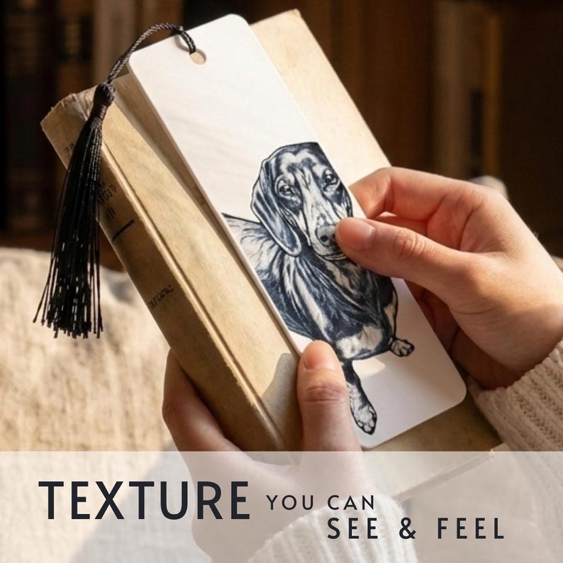 May include: A rectangular white bookmark featuring a detailed black and white ink illustration of a dachshund dog. The bookmark has a hole at the top with a black tassel attached. The text "TEXTURE YOU CAN SEE & FEEL" is printed at the bottom.