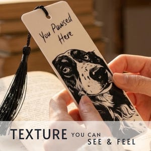 May include: A rectangular off-white bookmark featuring a black and white illustration of a dog's face and the text "You Pawsed Here" printed in black. The bookmark includes a hole at the top with an attached black decorative tassel.